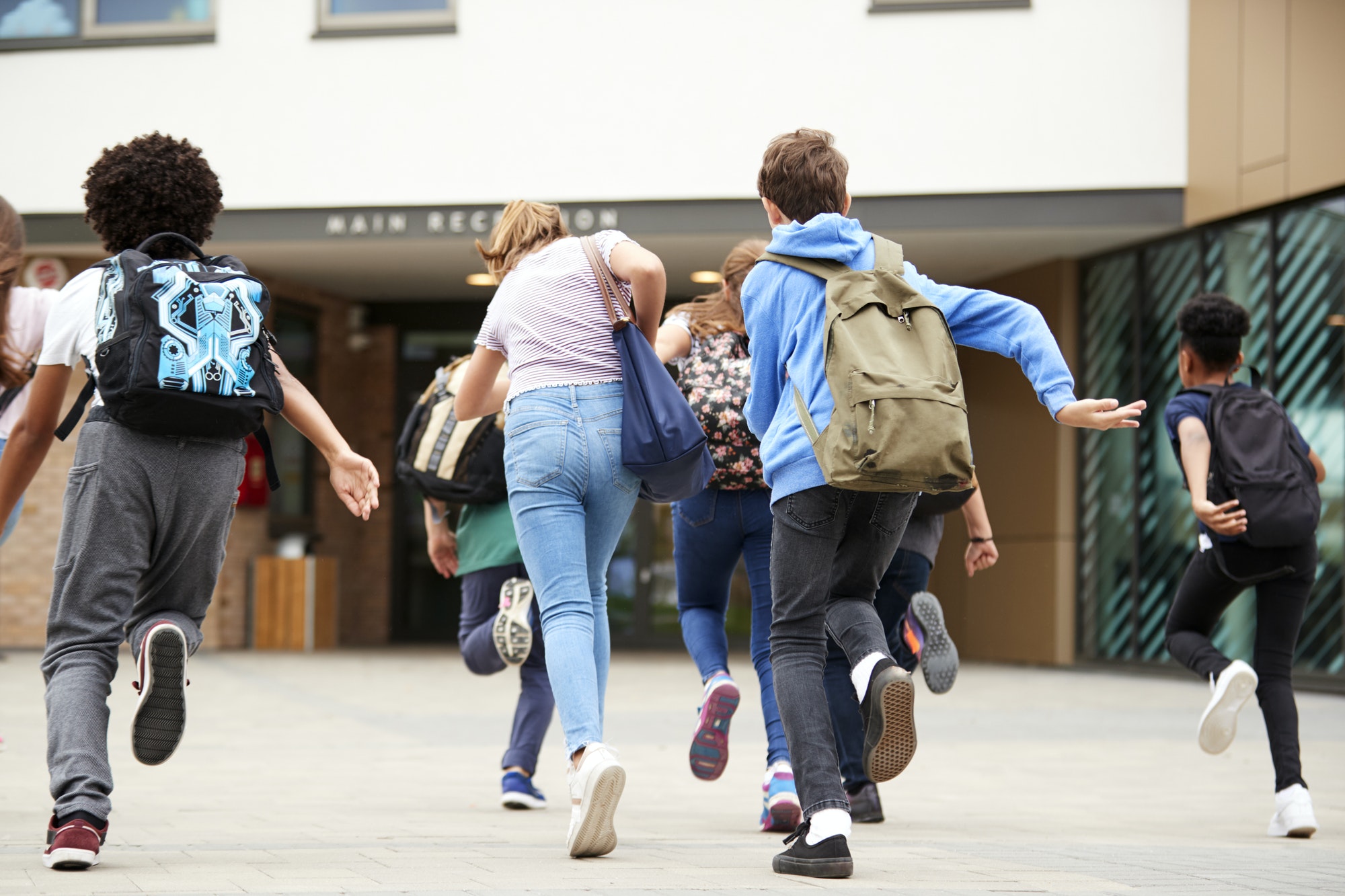 Group Of High School Students Running Into School Building At Beginning Of Class