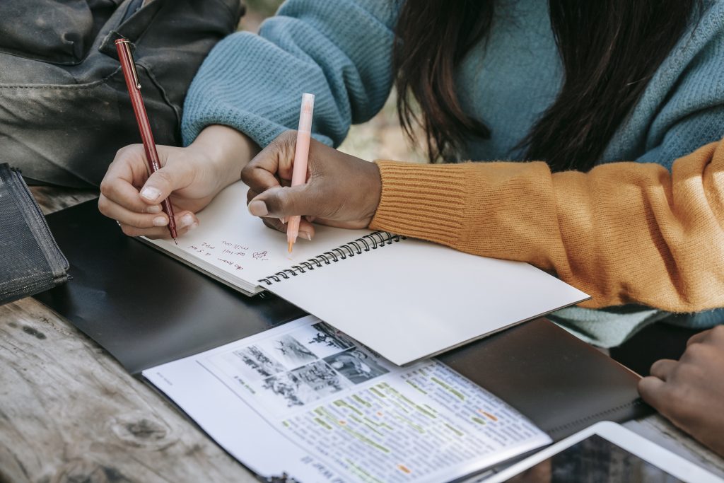 Crop unrecognizable diverse schoolgirls taking notes in copybook in park